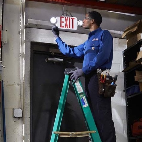 A Cintas technician standing on a ladder inspecting an emergency exit light fixture above a black door in a storage area.
