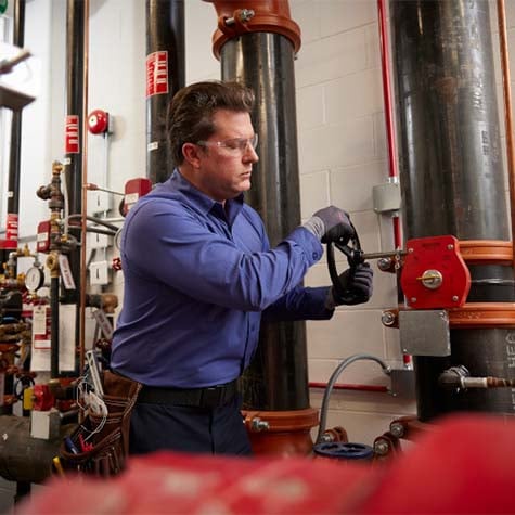A Fire Service Technician adjusting a sprinkler riser in a mechanical room with various valves and pipes.